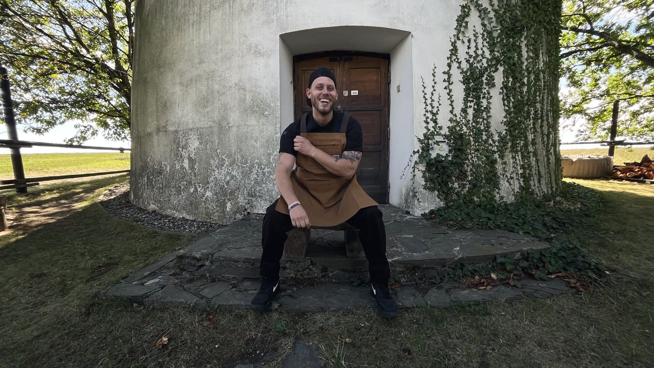 A cook in a leather apron sitting on the stone steps of a round whitewashed building, ivy on the wall behind him