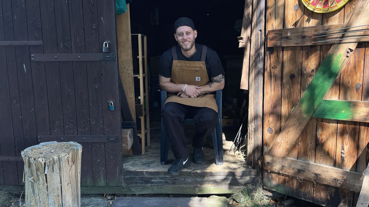 A cook in a black cap and tan apron sitting on the wooden steps of a shed, its doors open behind him