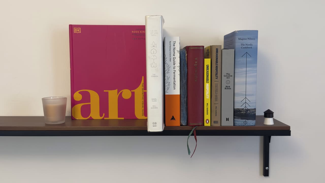 A wall shelf of cookbooks and reference held between a candle and a small lamp, spines out — Art, the Noma fermentation guide, Unreasonable Hospitality, The Creative Act, The Nordic Cookbook