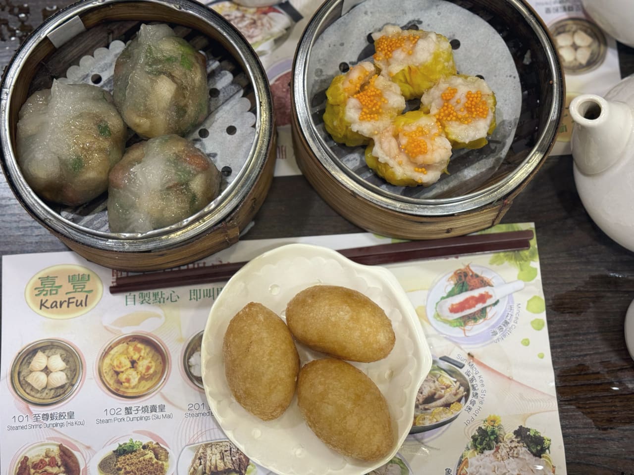 Two bamboo steamers of dim sum — translucent dumplings, and prawn siu mai topped with roe — beside three fried dumplings on a saucer, on a printed menu mat