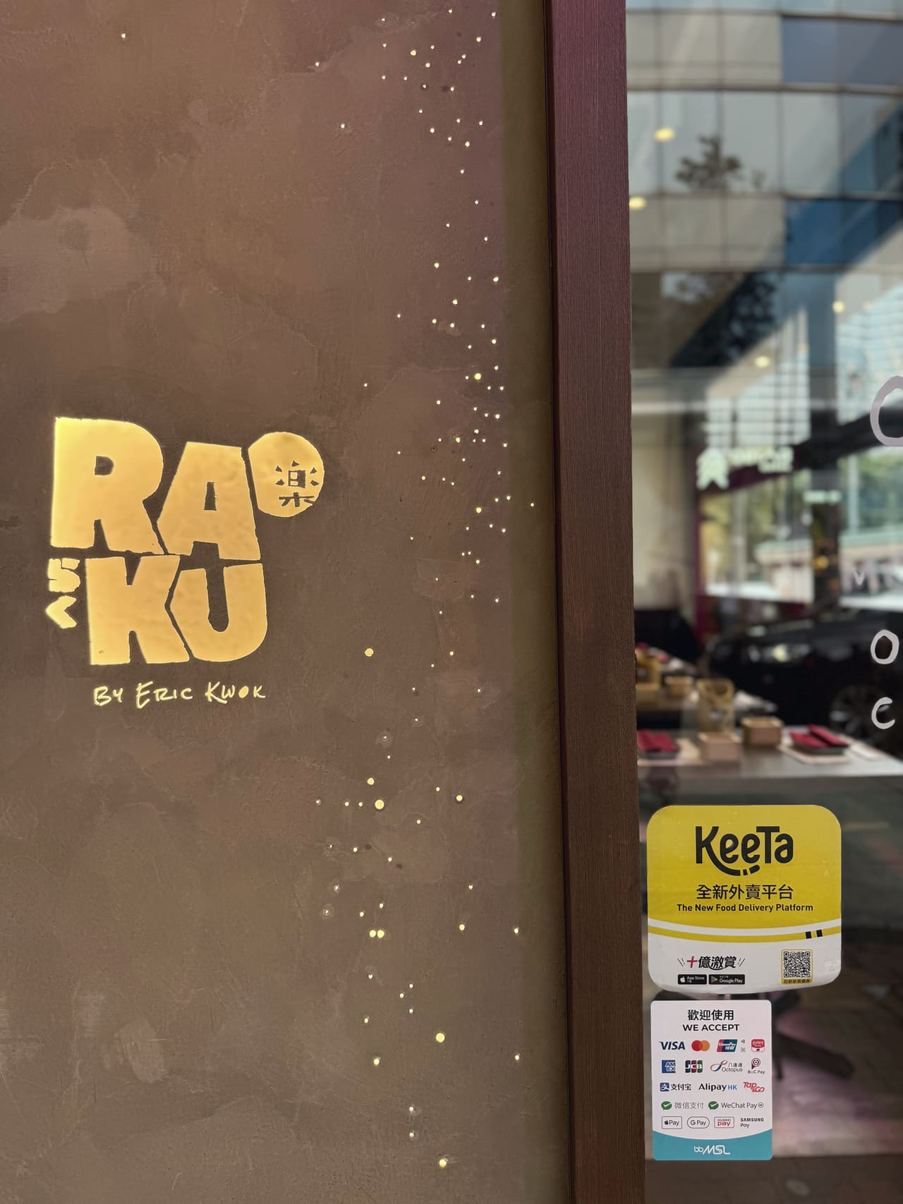 A restaurant doorway at street level, the name RAKU cut into a gold-lit panel beside the window