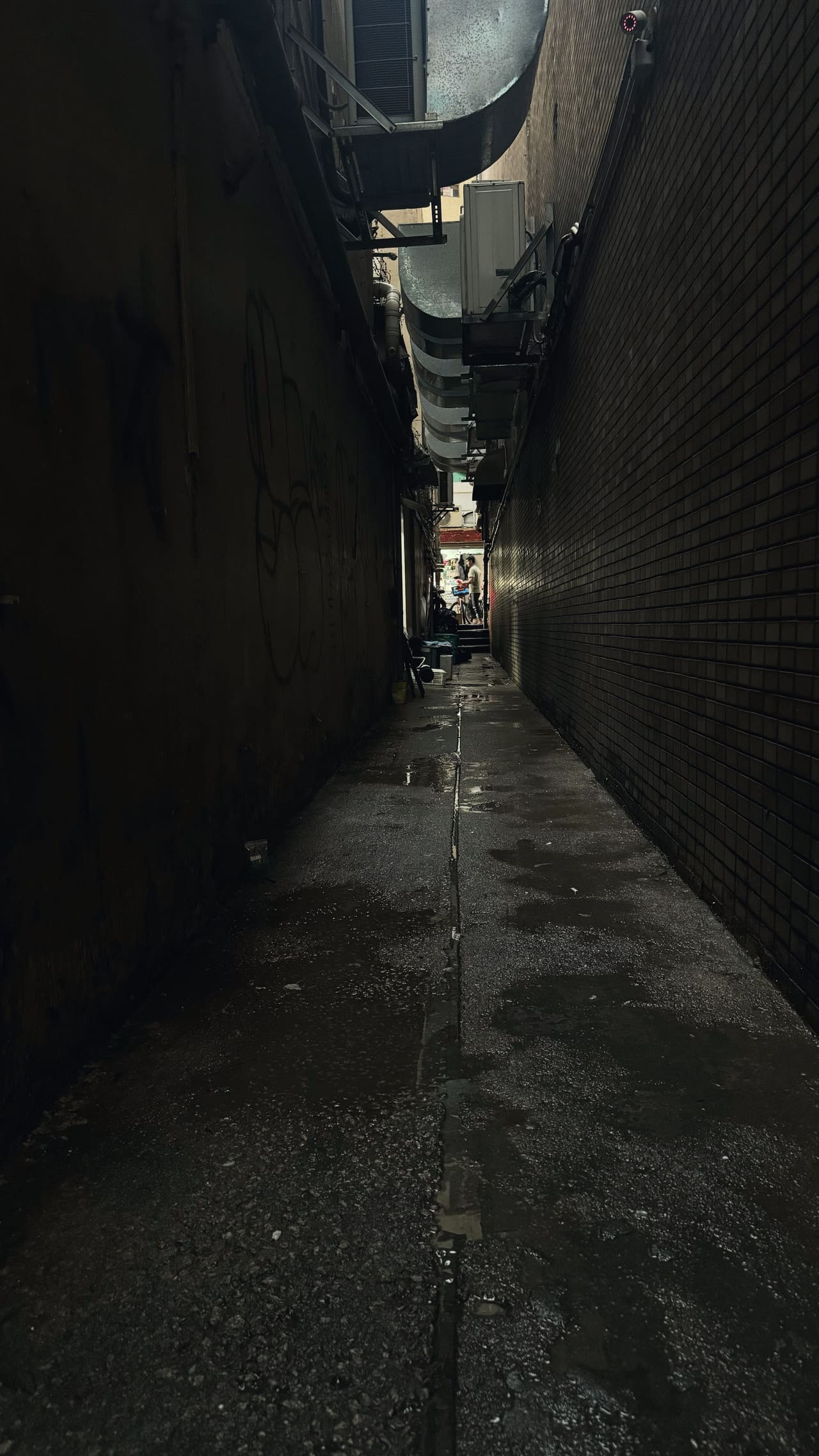 A narrow alley between two buildings, wet underfoot, air-conditioning ducts running overhead, a lit street at the far end