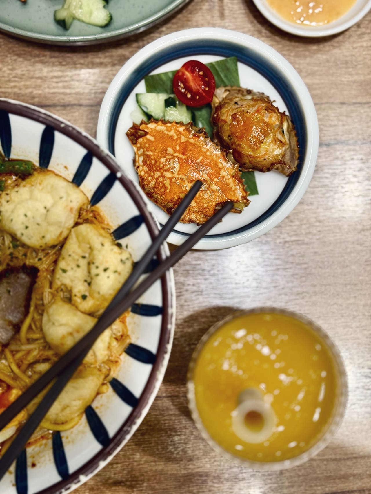A bowl of noodles in chilli broth topped with dumplings, a side plate holding a stuffed crab shell and a piece of roast meat, and a glass of mango drink