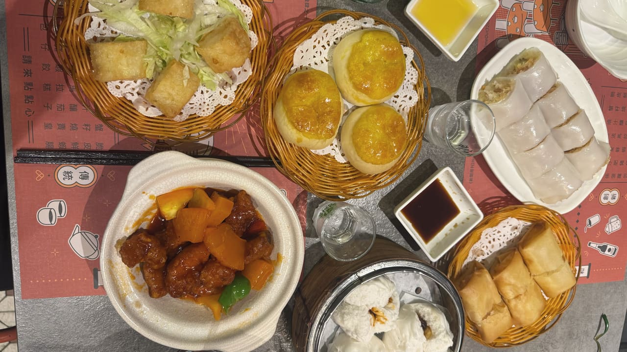 A table of dim sum seen from above — fried tofu, glazed buns, rice noodle rolls, sweet and sour pork, steamed buns and spring rolls