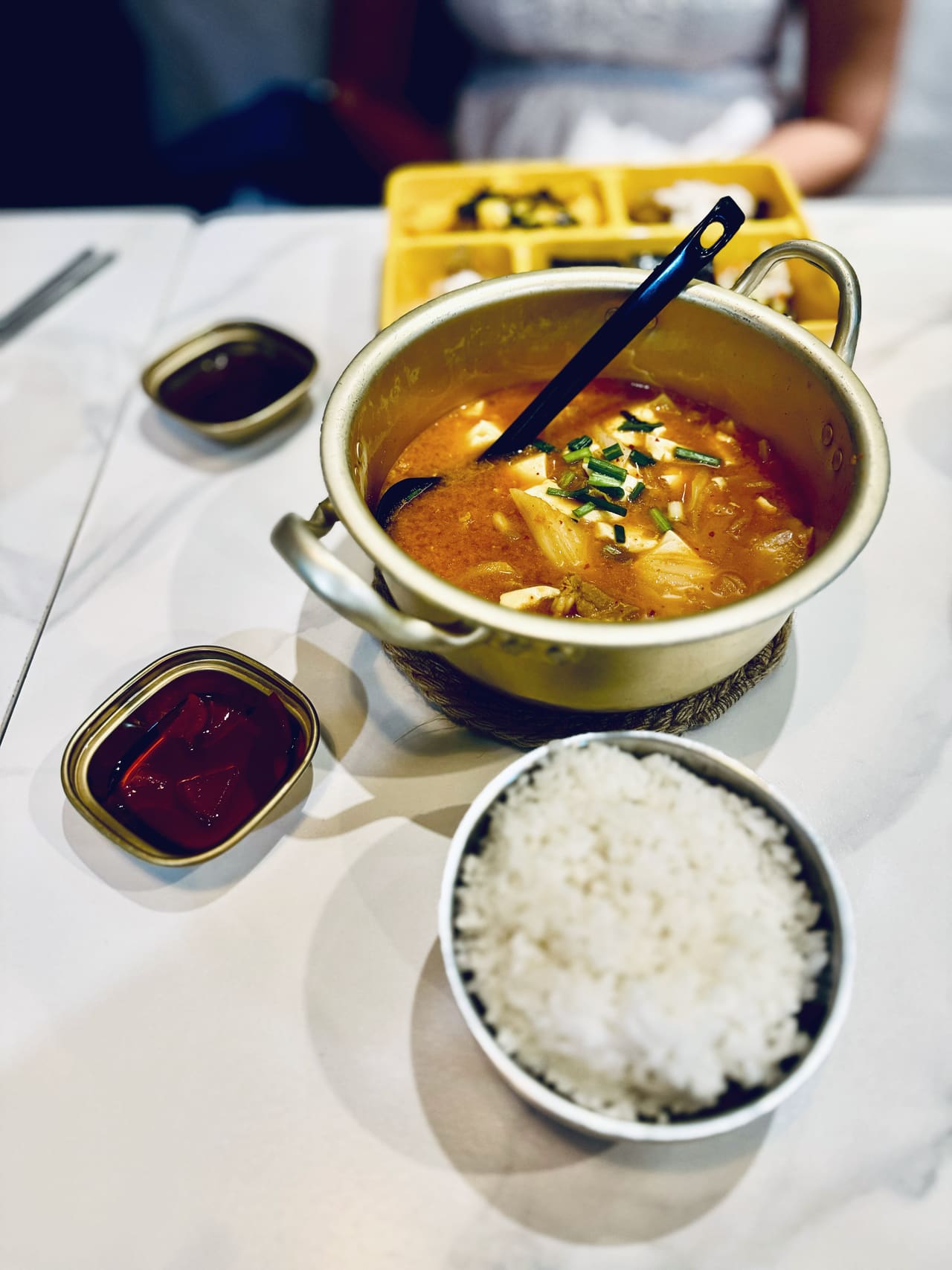 A brass pot of kimchi stew on a white cloth, a bowl of white rice and small dishes of pickles beside it