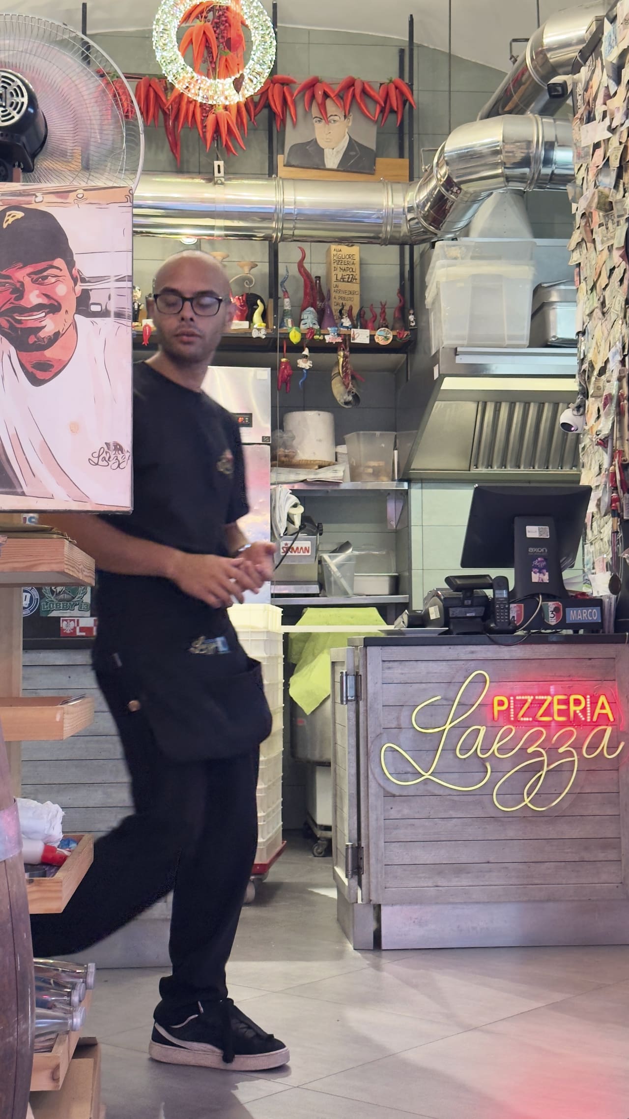 Inside a pizzeria — a cook crossing the room in black, strings of red chillies hung over the counter, a neon sign reading Pizzeria Laezza