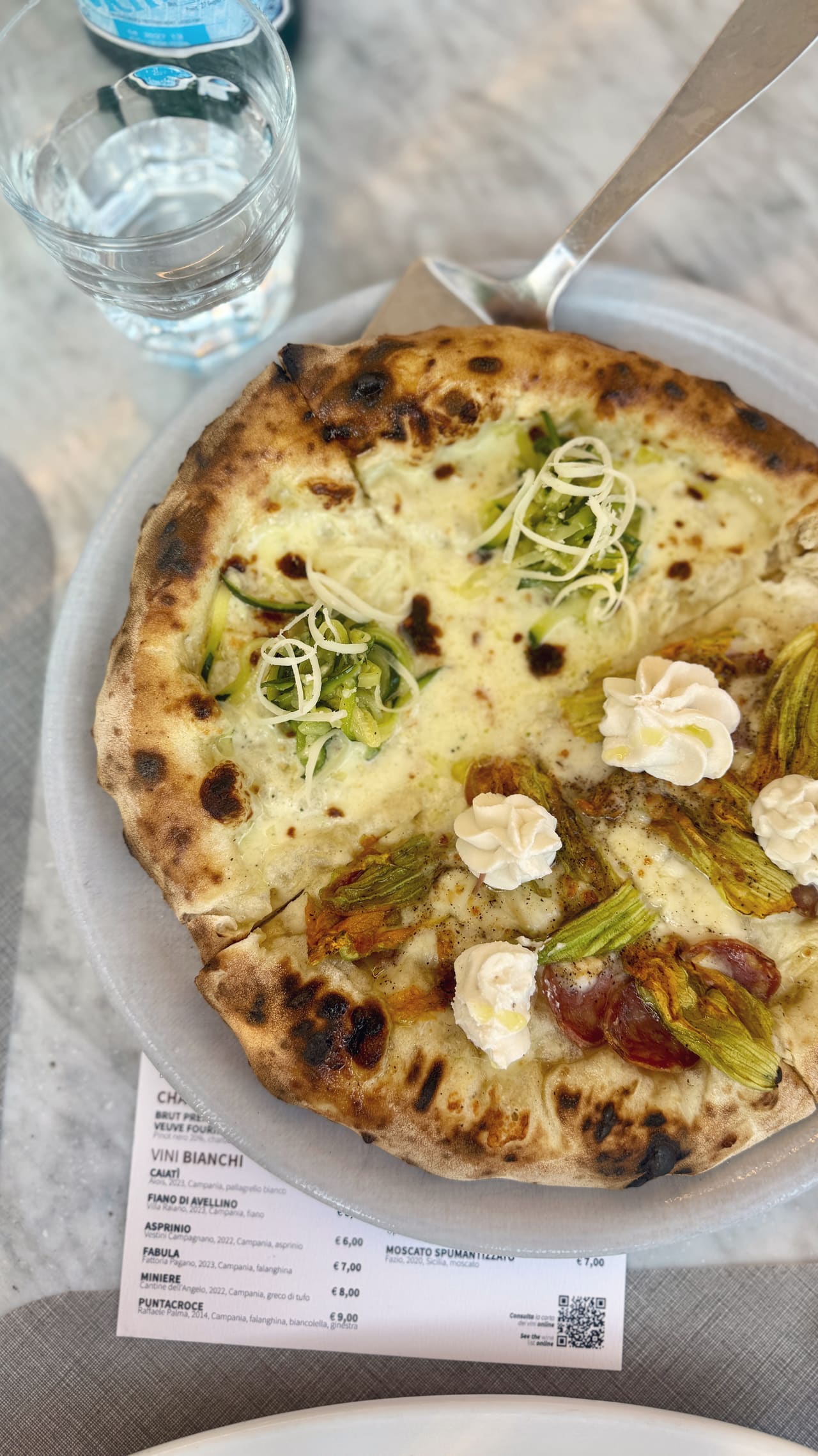 A white pizza with courgette, shaved cheese, ricotta and courgette flowers, the wine list lying under the plate