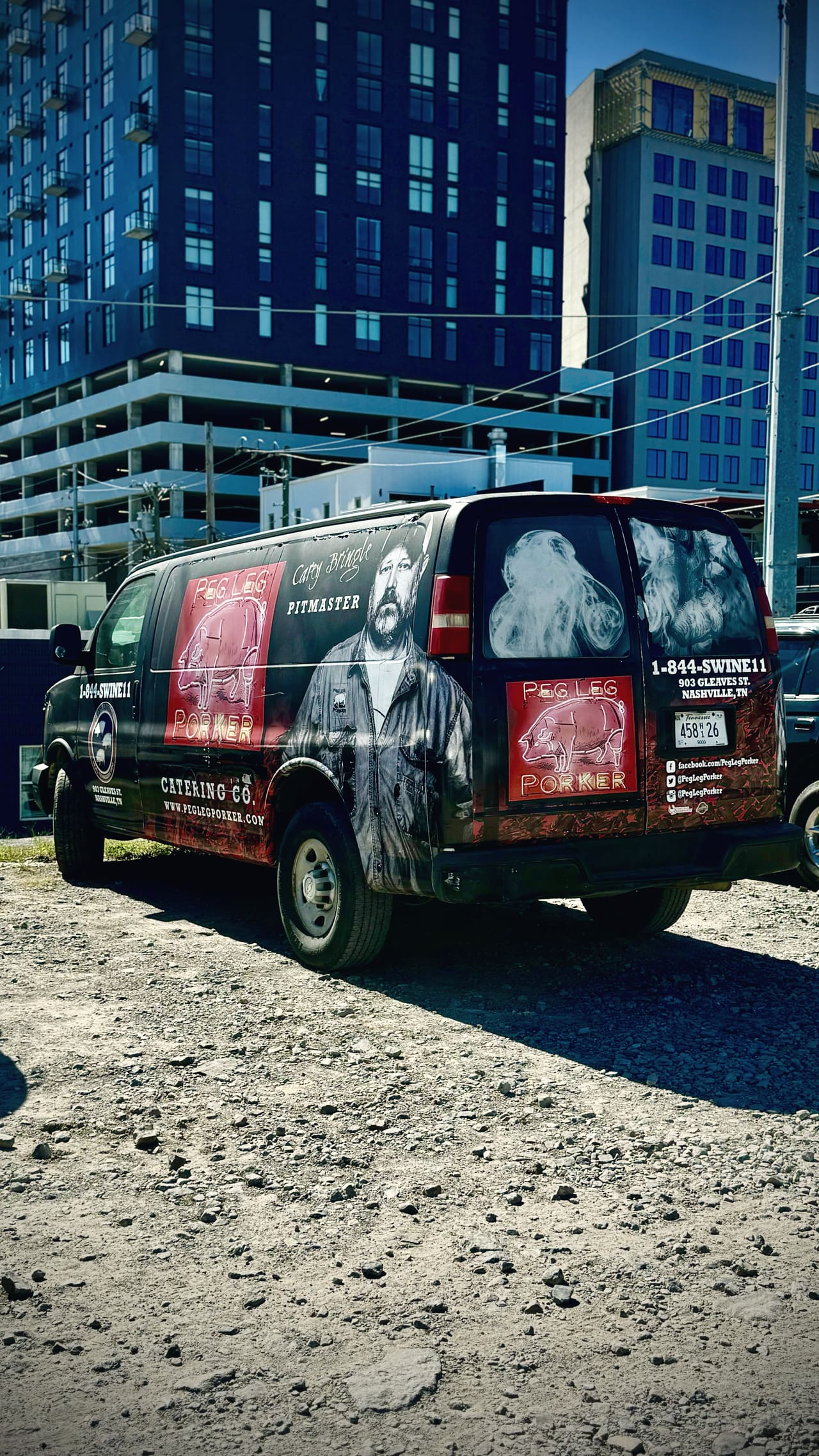 A black catering van parked on gravel, a pitmaster’s portrait and the words Peg Leg Porker painted down its side, glass towers behind