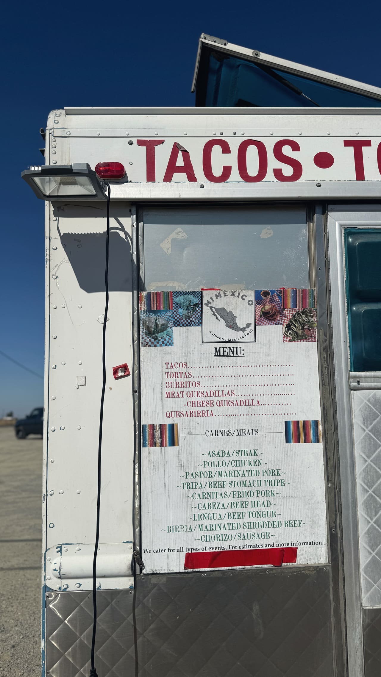 The service window of a taco trailer against a bare blue sky, its handwritten menu listing asada, carnitas, lengua, cabeza, birria