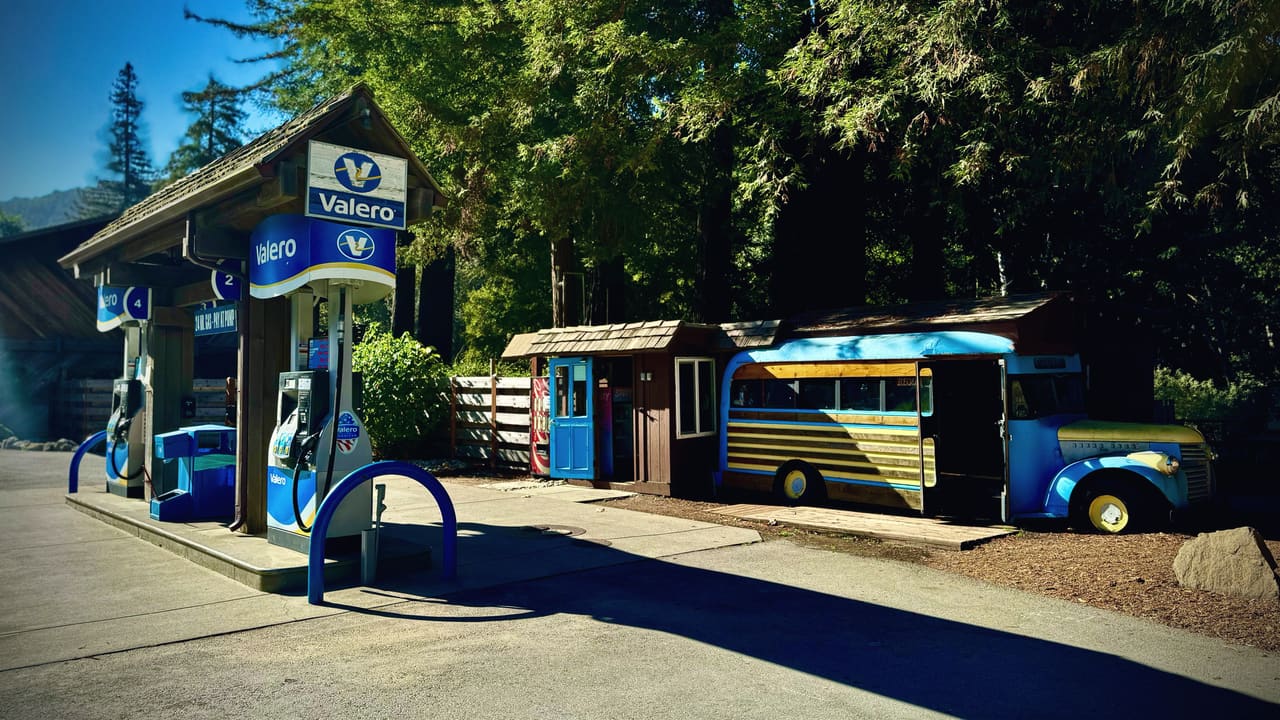 A roadside filling station under redwoods, an old blue school bus parked alongside it with its door open